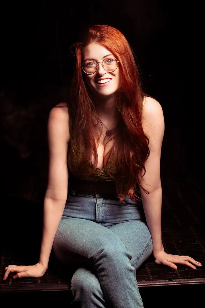 Fiona Cauley smiles at the camera. She has long, straight red hair and round wire-rimmed glasses, and wears a dark olive top and light-wash jeans. She sits on the edge of a metal platform against a black background.